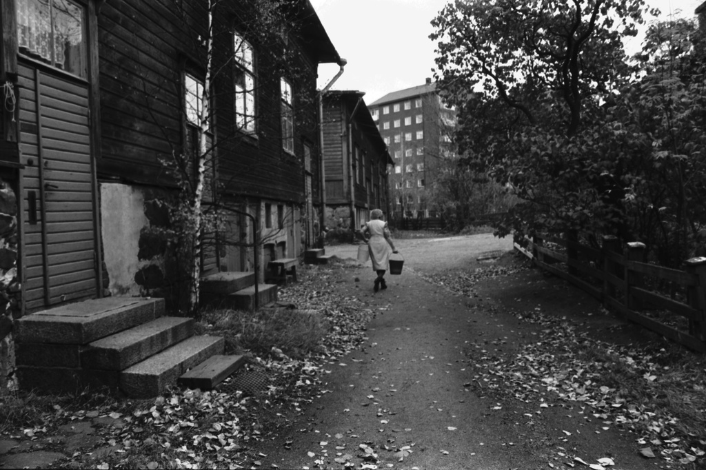 Lastenkodinkatu 2-10. Ruoholahden sadan markan villat. Nainen ämpärien kanssa puutalojen syksyisessä pihapiirissä. Taustalla Ruoholahdenkatu 18:n kerrostalo.