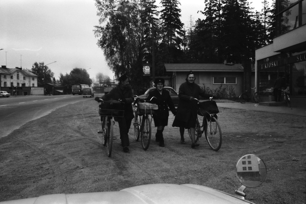 Malmi Ylä-Malmi. Kolme naista, todennäköisesti postinjakajia, polkupyörineen Kirkonkyläntiellä. Syksyinen näkymä auton ikkunasta. Oikella talo, jossa ravintola Malmi Kesti ja R-kioski. Kioskin luukulla mies asiomassa.