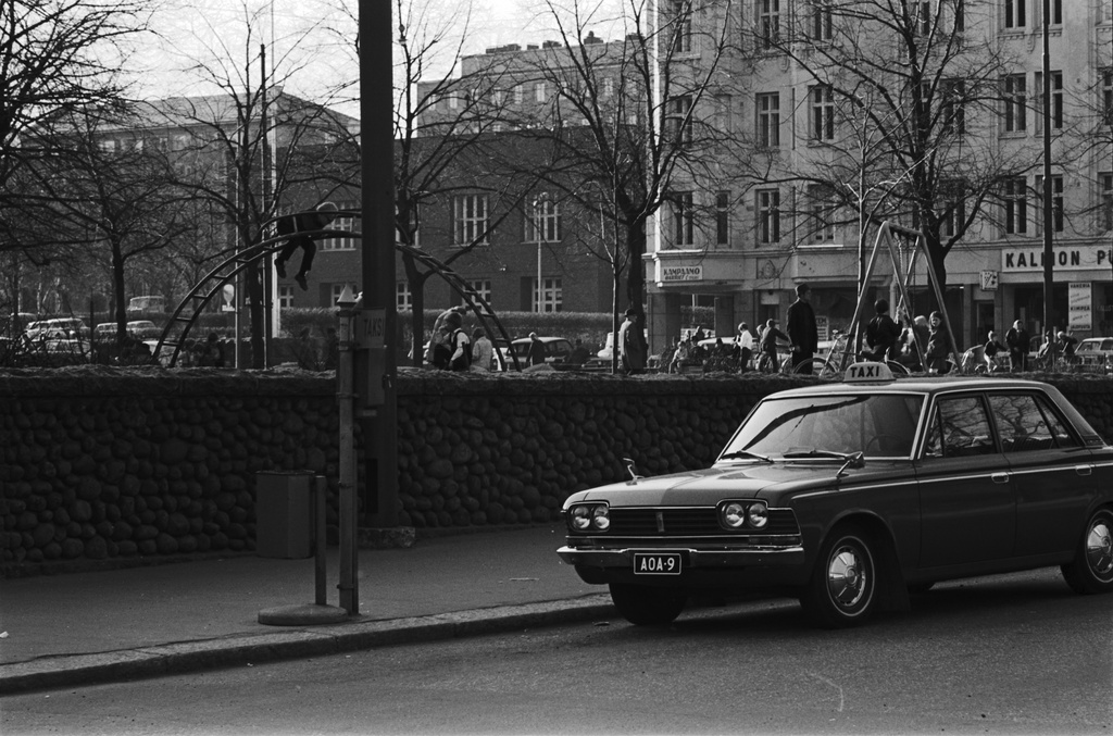 Karhupuisto. Fleminginkatu. Taksi tolpalla Fleminginkadulla Karhupuiston kiviaidan laidalla Viidennen linjan kulmassa. Puistossa lapsia ja aikuisia. Näkymä Agricolankadun suuntaan, vasemmalla Kallion paloasema (nykyinen Keskuspelastusasema). Auto oikealla: Toyota Crown. Agricolankatu 15, oikealla Agricolankatu 13.