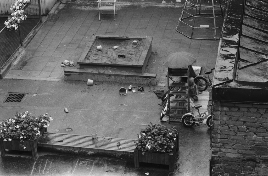 Liisankatu 7. Näkymä sadesäällä Liisankatu 7:n neljännen kerroksen ikkunasta pihan perällä olevalle lasten leikkipaikalle. Lapsia leikkimässä liukumäen alle rakentamassaan majassa. Keskellä pieni hiekkalaatikko.