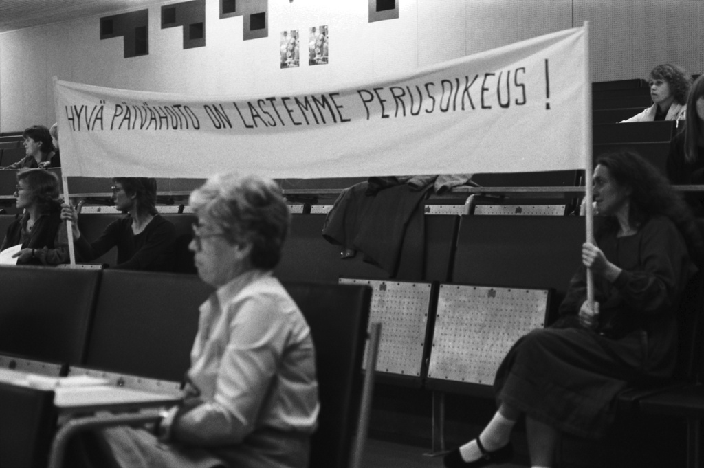Yliopistonkatu 3. Lasten päivähoito-oikeuksia käsittelevä tilaisuus Helsingin yliopiston Porthanian suuressa luentosalissa Hallituskatu 7:ssa (Yliopistonkatu 3). Lehtereillä istuvien naisten pitelemässä banderollissa teksti: Hyvä päivähoito on lastemme perusoikeus!
