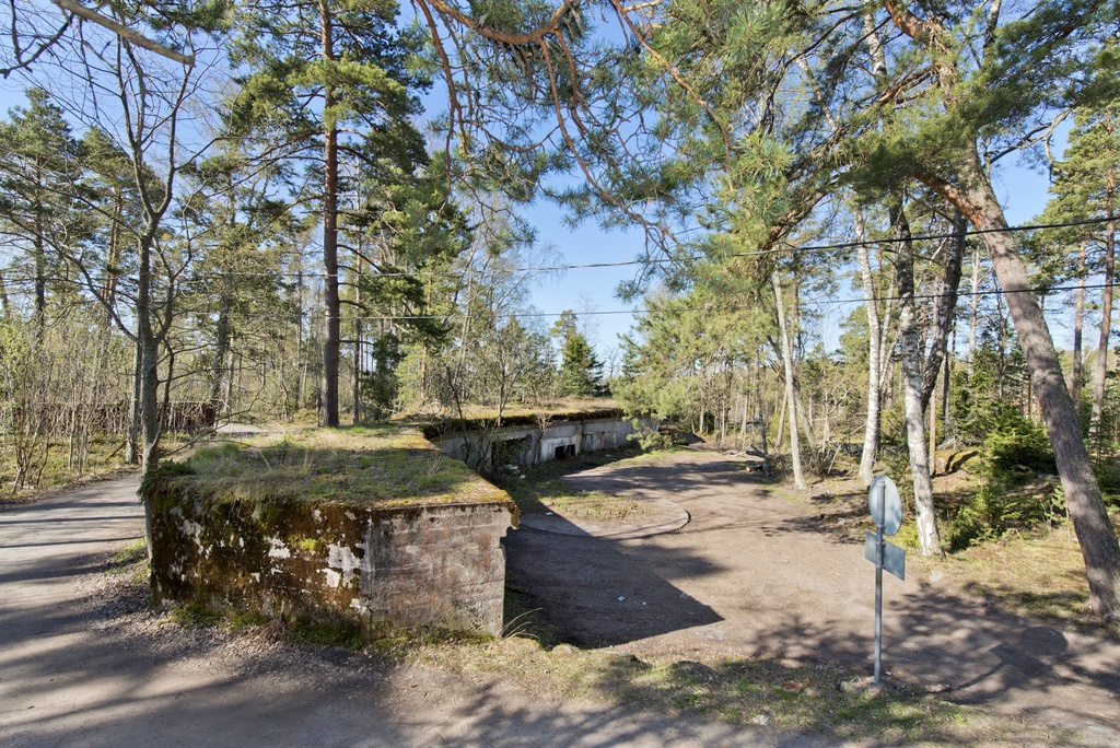 Skatanniemi. Ensimmäisen maailmansodan aikainen rannikkotykkipatteri Skatanniemessä, Vuosaaressa. Kuvassa on rintasuoja, jonka taakse on sijoitettu yhteensä neljä tykkiasemaa. Betonista valmistetun rintasuojan seinässä näkyy ampumatarvikekomero. Seinällä erottuu myös kaksi pientä luukkua, joista näkee sisälle rintasuojan alle rakennettuihin miehistö- ja varastotiloihin. Keskellä on betonista valettu jalusta itse tykille, jota ei koskaan ehditty asentaa. Jalustan keskellä oleva pulttirinki kertoo, mihin tykki oli tarkoitus kiinnittää. Etualalla oikealla on liikennemerkki. Kuvassa vasemmalla aseman edestä ja läpi kulkee tie, joka on puhkaistu asemaan 1920-luvulla. kuvaussuuntana lounas.