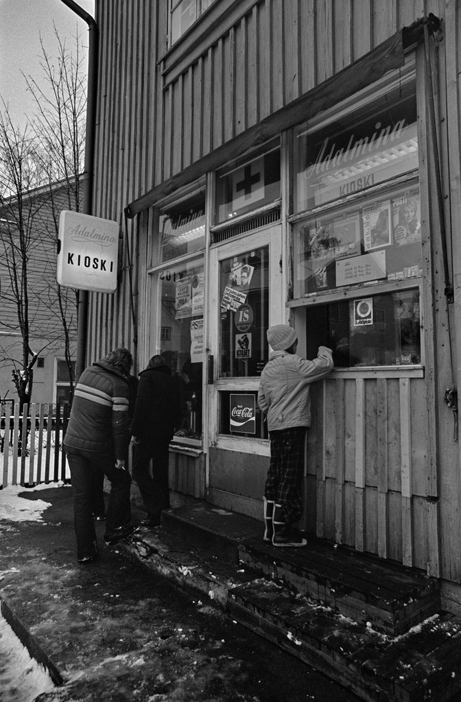 Kioski Adalmina puurakennuksessa Toukolassa. Kioskin ikkunoihin on kiinnitetty mainoksia ja lehtien lööppejä. Kioskin palveluluukun yläpuolella roikkuu myytäviä lehtiä. Lapsiasiakas on asioimassa palveluluukulla.