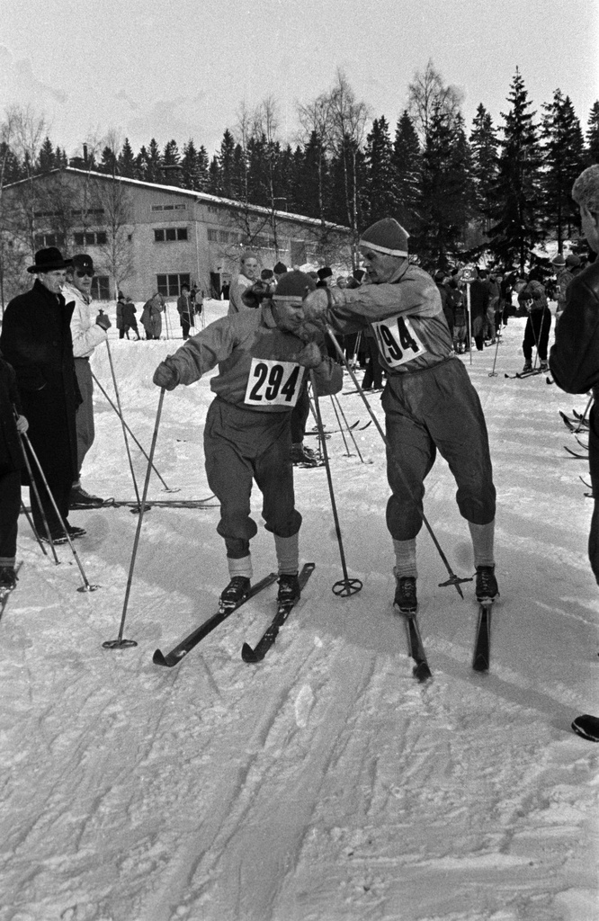 Hiihtokilpailut Ruskeasuon liikuntapuistossa. Hiihtäjä antaa toiselle kaulassa roikkuvaa viestikapulaa. Taustalla Ruskeasuon tallit.