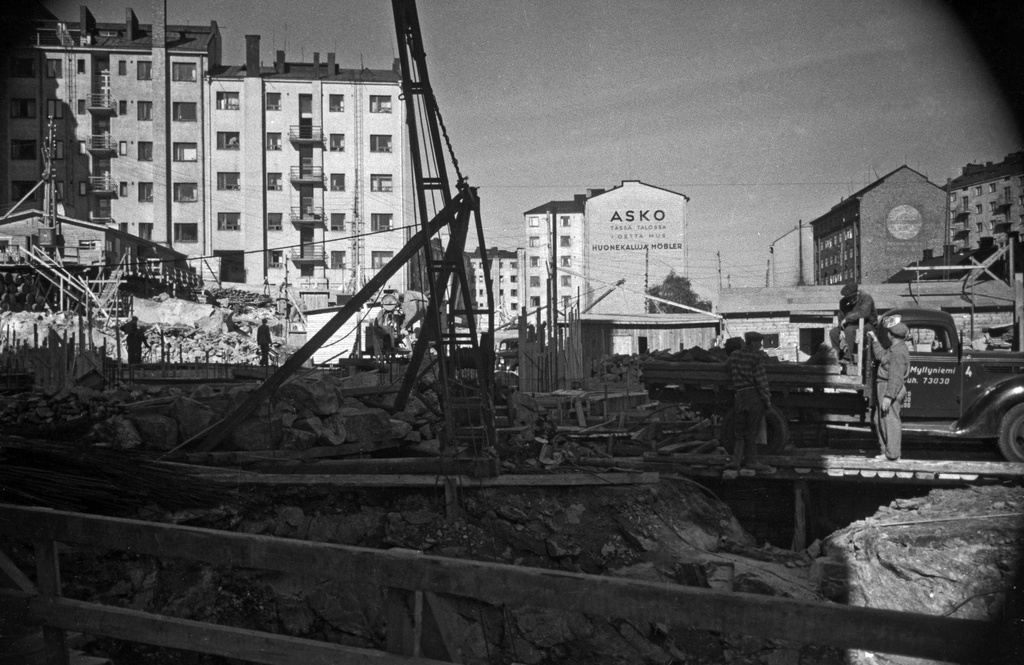 Rakennustyömaa Hämeentiellä, Hämeentie 44, 42 rakenteilla. Vasemmalla sisäpihalta nähtynä Torkkelinkatu 4, 2 b. Asko-liikettä mainostava talo on Hämeentie 48. Oikealla Hämeentie 23, jonka takana oikealla näkyy Vilhovuorenkuja 6.