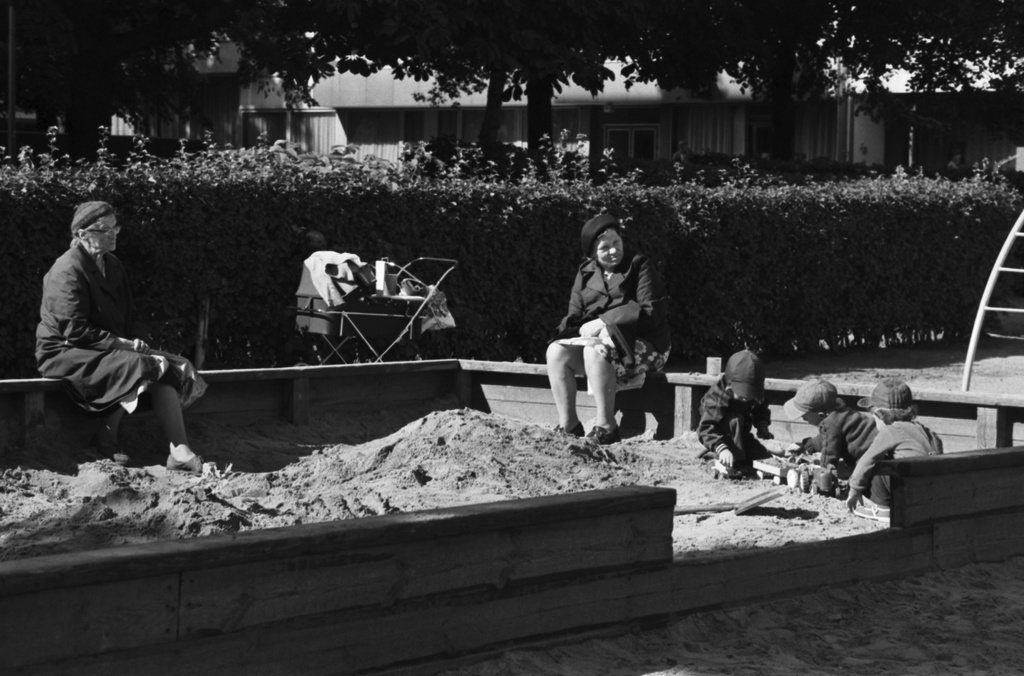 Tehtaanpuisto. Lapsia leikkimässä hiekkalaatikossa Tehtaanpuiston leikkipaikalla. Laatikon reunalla istumassa kaksi iäkästä naista.