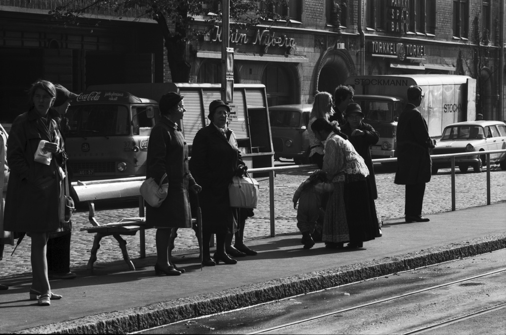 Mannerheimintie 1. Ihmisiä, oikealla romaninainen pienen lapsen kanssa, raitiovaunupysäkillä Mannerheimintiellä Stockmannin tavaratalon edustalla.