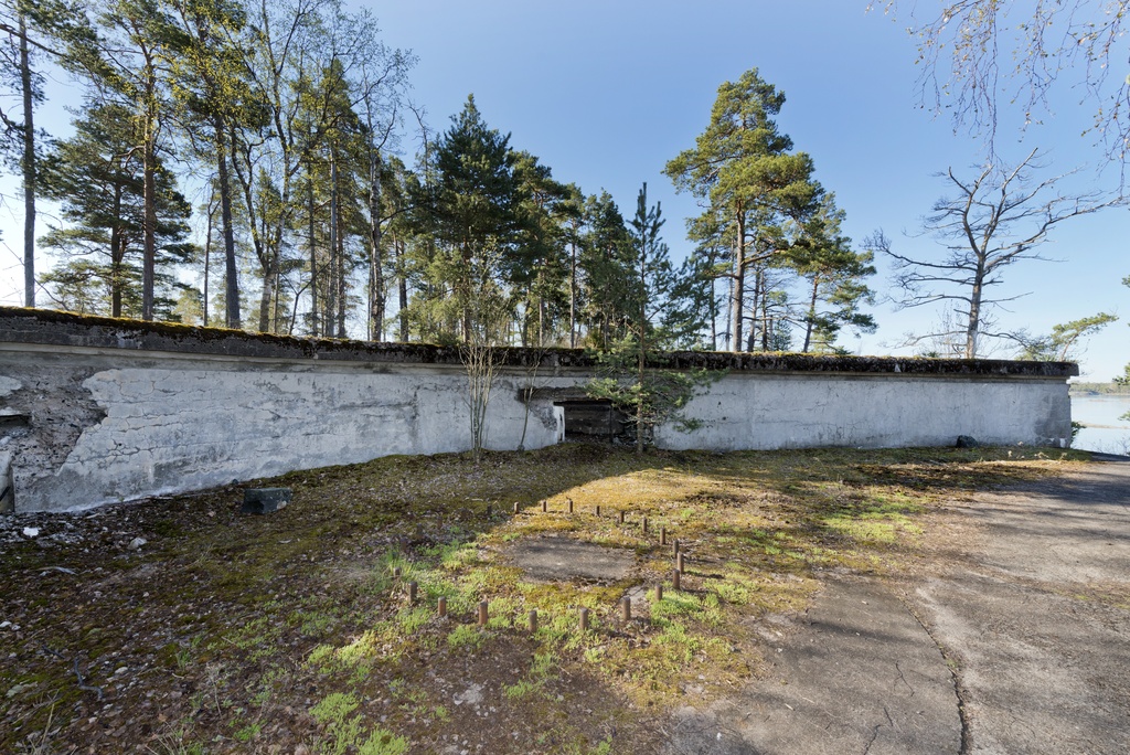 Skatanniemi. Ensimmäisen maailmansodan aikainen rannikkotykkipatteri Skatanniemessä, Vuosaaressa. Kuvassa on rintasuoja, jonka taakse on sijoitettu yhteensä neljä tykkiasemaa. Betonista valmistetun rintasuojan seinässä näkyy ampumatarvikekomero, jonka edessä kasvaa pieni mänty. Rintasuojan taustalla on betonista valettu pyöreä jalusta itse tykille, jota ei koskaan ehditty asentaa. Jalustan keskellä oleva pulttirinki kertoo, mihin tykki oli tarkoitus kiinnittää. Kuvaussuuntana etelä.