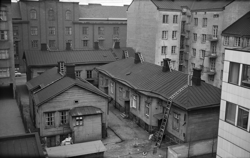 Valokuva sisäpihalta, missä monikerroksisten kivirakennusten ympäröiminä on kolme toisiaan lähelle rakennettua yksikerroksista puusta rakennettuja asuintaloa, jotka puolestaan muodostavat keskelleen pienen piha-alueen. Tuon piha-alueen kahden puolen olevista taloista toisesta näkyy kolme sisäänkäyntiä pihan puolelta. Taustalla näkyy Teknillisen korkeakoulun rakennus tien toiselta puolen (Eerikinkatu 36).