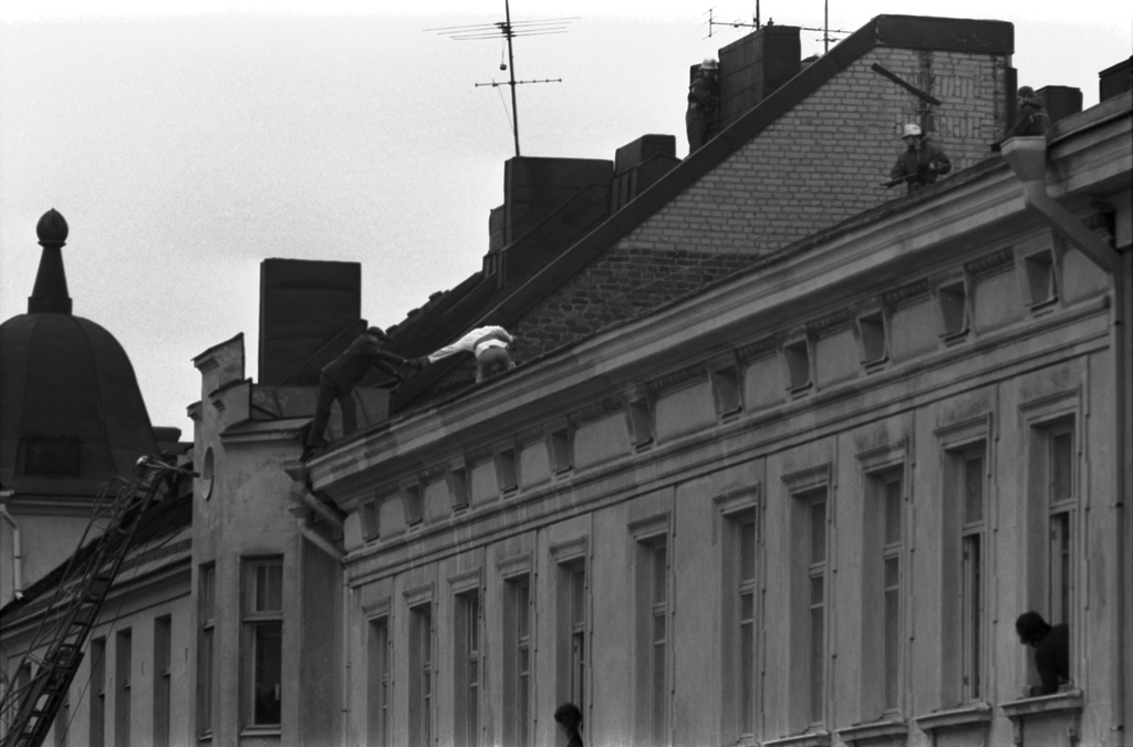 Meritullinkatu 22, 20. Meritullinkatu 22:n katolla oleva pelastusmies ojentanut kätensä Meritullinkatu 20:n katolle kiivenneelle miehelle. Meritullinkatu 22:n ikkunoista kurkistelee ihmisiä seuraten tapahtumaa.
