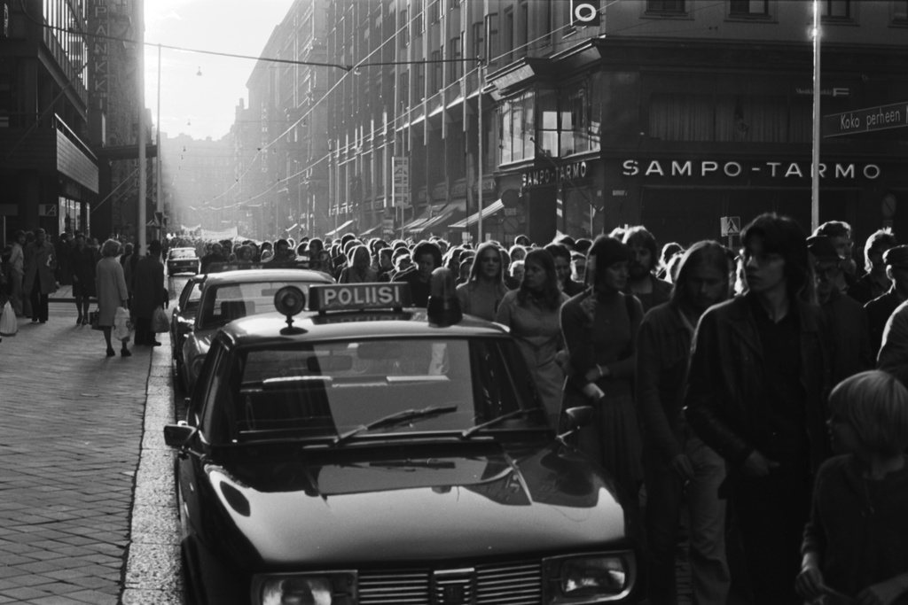 Aleksanterinkatu. Chilen sotilasjuntan vastainen mielenosoituskulkue iltapäivän tai alkuillan auringossa Aleksanterinkadulla. Etualalla kadun laidalla poliisiauto. Oikealla Sampo-Tarmon konttori Kluuvikadun kulmassa. Näkymä länteen Mannerheimintien suuntaan.