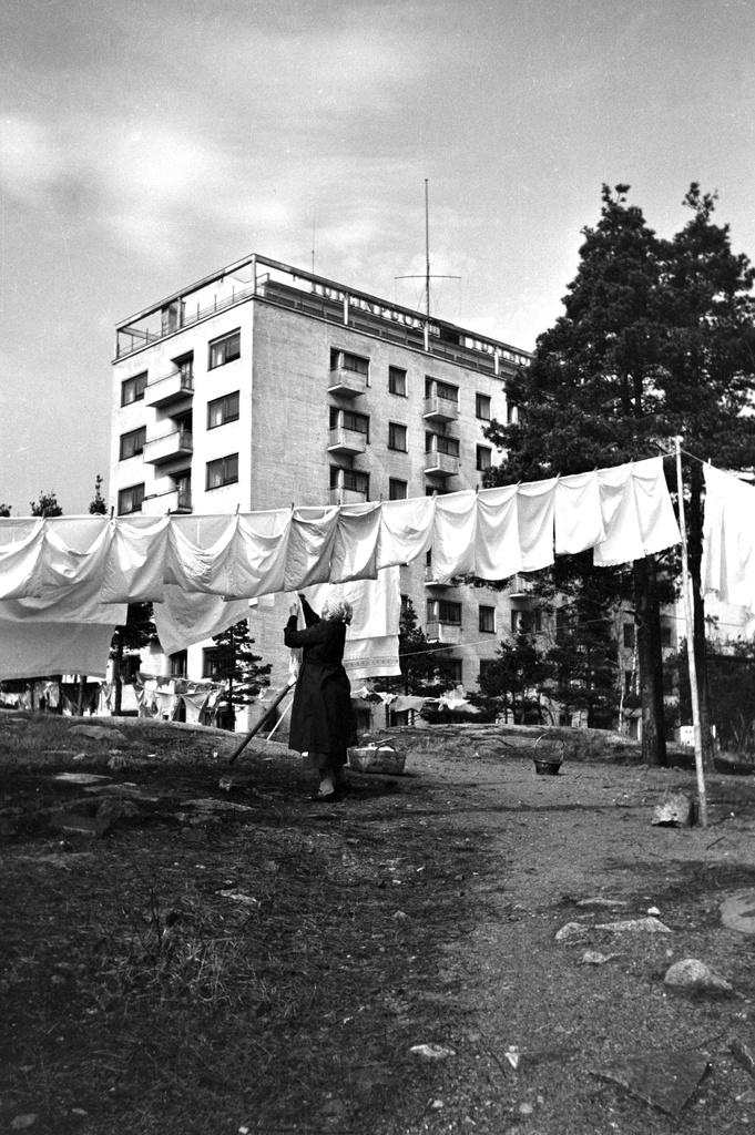 Nainen ripustaa pyykkejä narulle kuivumaan. Taustalla Mannerheimintie 118.