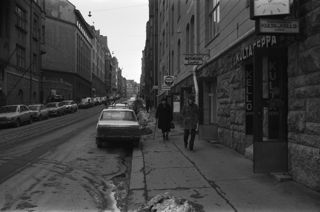 Katajanokka, Luotsikatu. Näkymä Satamakadun suunnalta Luotsikatu 2:n edestä itään Vyökadulle päin. Oikealla rivi kivijalkamyymälöiden avoimia ovia, ensimmäisenä Katajanokan Kulta- ja Kelloliike. Taaempana Parturiliike Luotsi, kampaamo, sekatavarakauppa sekä kenkäkauppa.