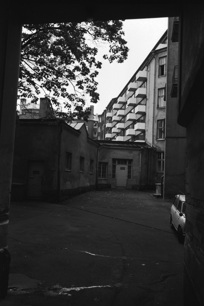 Pursimiehenkatu 27. Näkymä porttikäytävästä Pursimiehenkatu 27:n pihalle, jossa matala piharakennus. (Rakennus on entinen hevostalli ja puusepänverstas, joka vuonna 1996 remontoitiin asuinrakennukseksi.) Asfalttipiha. Taustalla Pursimiehenkatu 25:n kerrostalo parvekkeineen.