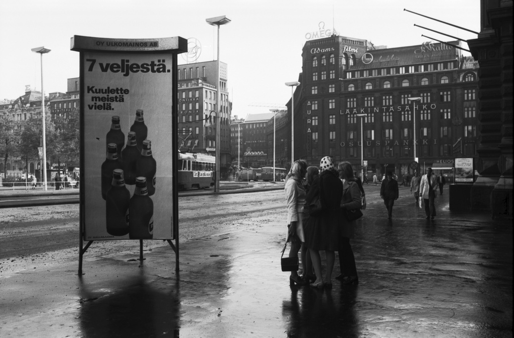 Kaivokatu. Mikonkatu 13. Näkymä Kaivokadulta Ateneumin edustalta Kaisaniemenkadun suuntaan. Vasemmalla Mallasjuoman 7 veljestä -olutmainos Oy Ulkomainos Ab:n mainospylväässä, oikealla neljän hengen naisryhmä seisomassa sateen kastelemalla kadulla. Taustalla oikealla Mikonkatu 13.