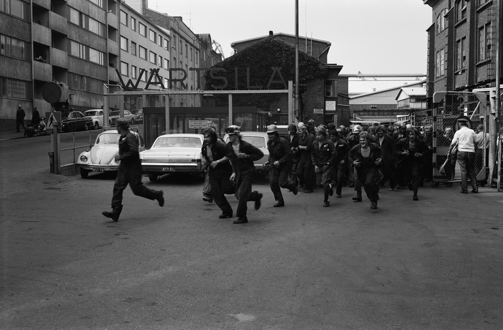 Wärtsilän telakan työntekijöitä menossa ruokatunnille, Länsisatama, kesä 1975. Telakkakatu. Vasemmalla Munkkisaarenkatu 4, 6, 8. Ajoneuvot Wärtsilä-merkin alla vasemmalta lukien: Volkswagen, Pontiac Tempest vm. 1965, Ford Taunus 17M. Kuvan vasemmassa reunassa pysäköidyt autot edestä lukien: Dodge Dart vm. 1965, Volvo, Ford Taunus, Volkswagen.