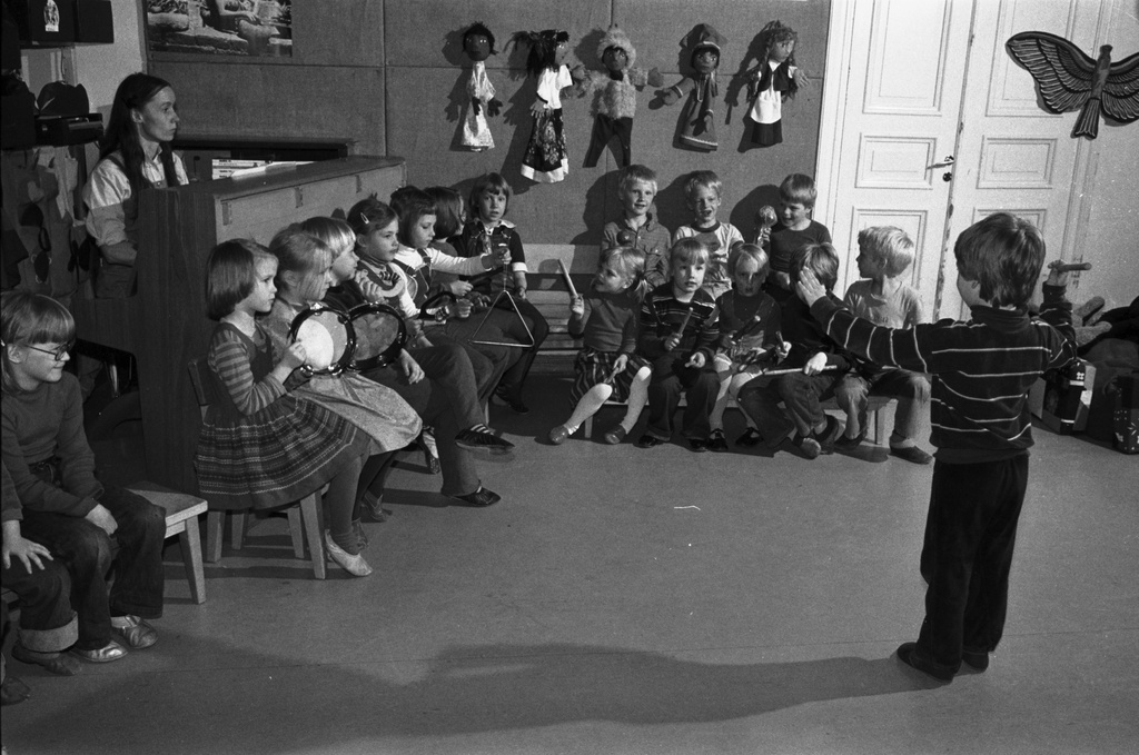 Rauhankatu 13. Lasten orkesteri esiintymässä Vironniemen päiväkodin salissa yhden lapsen toimiessa kapellimestarina. Lastentarhanopettaja säestää pianolla.