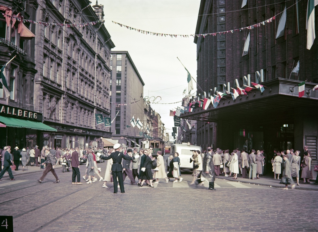 Helsingin olympialaiset 1952. Jalankulkijoita ja liikennettä Aleksanterinkadulla. Juhlaliputus Stockmannin tavaratalon pääsisäänkäynnin katoksella.