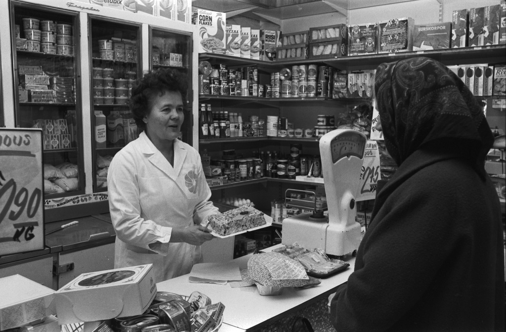 Liisankatu 7. Myymälänhoitaja ja asiakas Maanviljelijäin Maitokeskuksen myymälässä Liisankatu 7:ssä. Myyjä esittelee huivipäiselle asiakkaalle täytekakkua pienessä kivijalkamyymälässä. Myyjällä yllä valkoinen työtakki. Etualalla sardiineja ja muita kalasäilykkeitä.