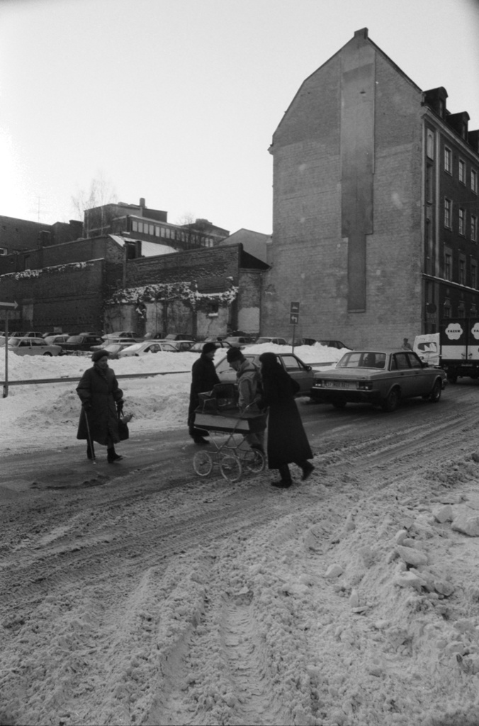 Kirkkokatu 4. Äiti (Etta Harvilahti) lastenvaunujen kanssa sekä muita ihmisiä ylittämässä lumista Kirkkokatua Meritullinkadun kulmassa. Taustalla oikealla Kirkkokatu 6:n talon päädyssä kuvataiteilijoiden Juha Sääsken, Raili Tangin ja Reijo Viljasen abstrakti seinämaalaus. Vasemmalla Kirkkokatu 4:n rakentamaton tontti.