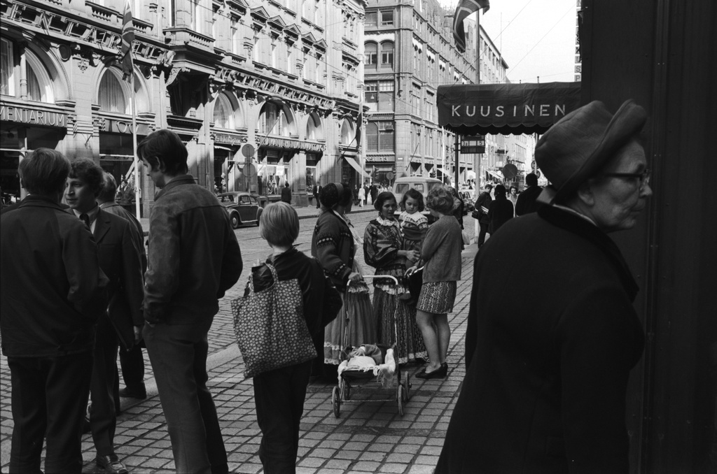Aleksanterinkatu. Näkymä Aleksanterinkatu 48:n edustalta Mikonkadun suuntaan. Etualalla oikealla nainen katselemassa näyteikkunaa. Vasemmalla ryhmä nuoria miehiä. Keskellä kaksi romaninaista lapsineen, oikealla sisäänkäynti Kuusisen vaateliikkeeseen. Taaempana jalankulkijoiden keskellä valokuvaaja Simo Rista kamerajalustoineen.