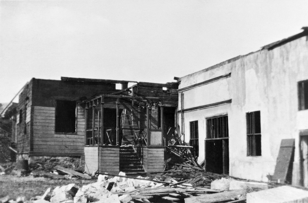 Näkymä purkutyön alla olevasta Eerikinkatu 30:n puurakennuksesta pihan puolelta. Seinät ovat vielä pystyssä mutta katto on kokonaan revitty ja kuistista on lähinnä kehykset jäljellä. Purkutyöt johtuvat samalle tontille vuonna 1925 valmistuneen Teknillisen korkeakoulun rakennustöiden käynnistymisestä. y Peltikatolla näkyy kolme savupiippua, räystästä vasten puiset tikkaat ja seinään on kiinni rakennettu puinen kuistimainen rakenne laudoista. Kuvan oikeassa reunassa näkyy pieni osa katoksellista rakennusta, jossa on kolmiomainen pääty. Valokuva on otettu kevättalven aikaan, jolloin maassa on vielä jonkin verran lunta. Iltapäivän aurinko paistaa kohtalaisen korkealta.