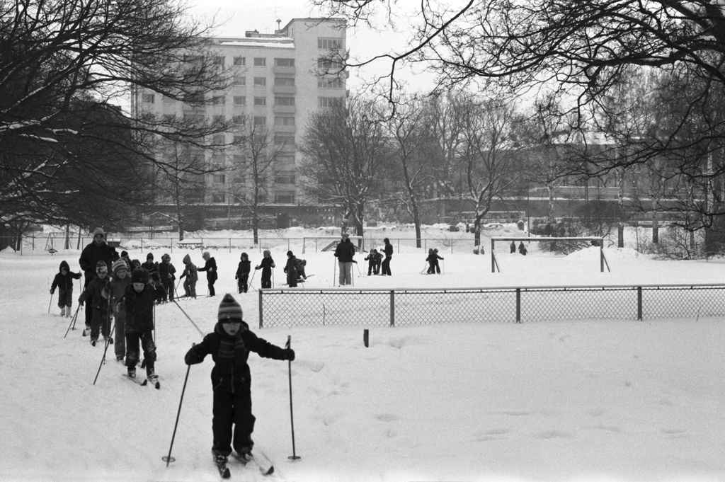 Kaisaniemenpuisto. Vironniemen päiväkodin lapset ja aikuiset lumisessa Kaisaniemenpuistossa lähdössä hiihtoretkelle. Taustalla Unioninkatu 39:n kerrostalo ja Unioninkatu 37:n kulma.
