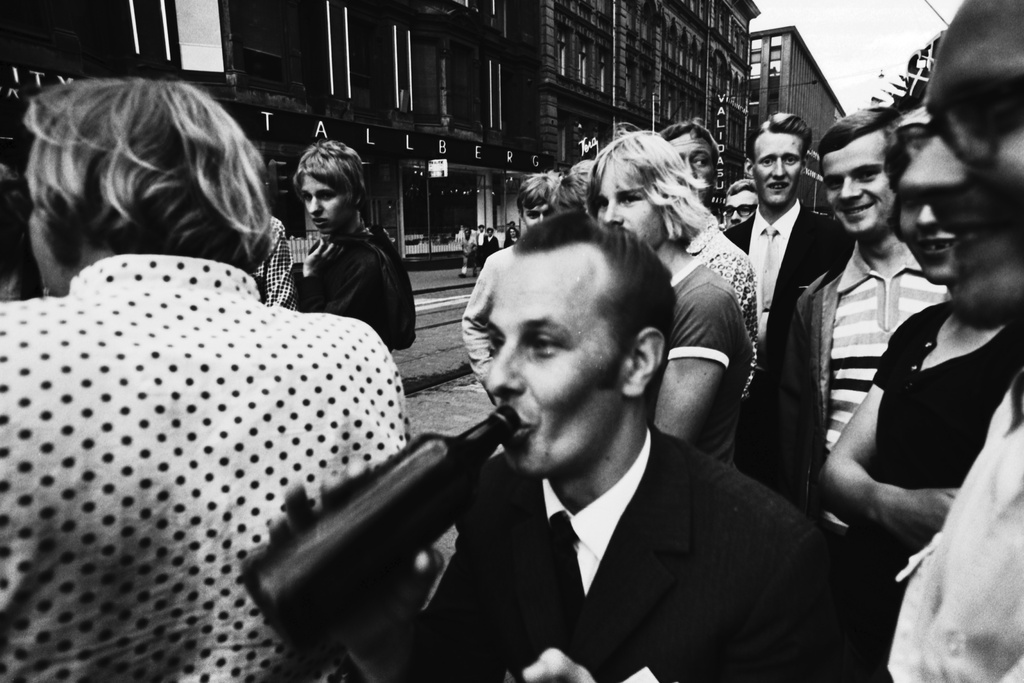 Aleksanterinkatu 52, 21. Mies juomassa pullon suusta iloisen ihmisjoukon ympäröimänä Stockmannin tavaratalon edustalla Aleksanterinkadun kävelykatukokeilun avajaispäivänä.