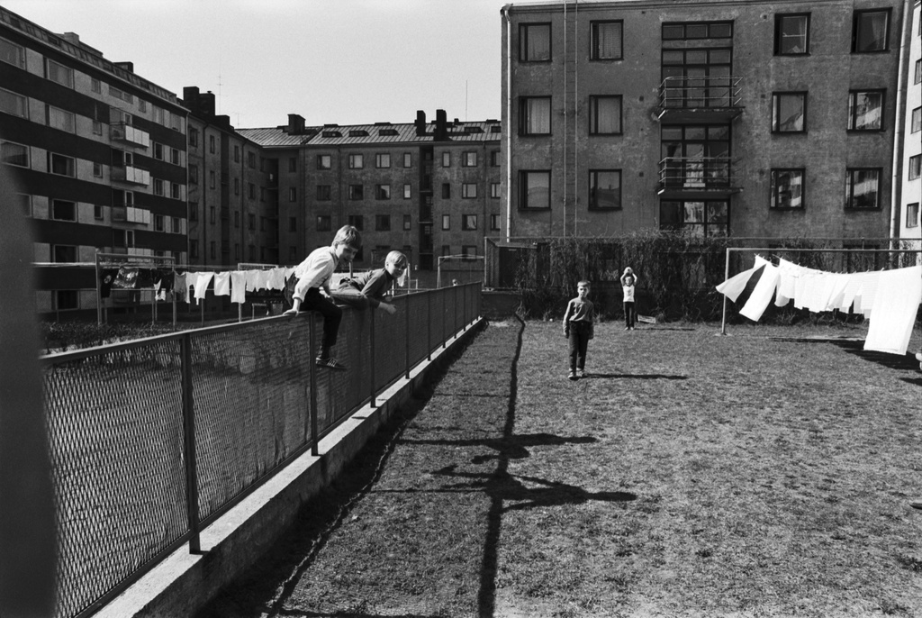 Neljäs linja 24. Näkymä Kolmas linja - Castréninkatu - Neljäs linja - Suonionkatu -katujen ympäröimän korttelin pihalta luoteeseen. Etualalla poikia kiipeämässä Kolmas linja 23 - Neljäs linja 24 välisen verkkoaidan yli sekä pelaamassa jalkapalloa valkopyykin liehuessa narulla kuivumassa. Oikealla Neljäs linja 26, taustalla vasemmalla Castréninkatu 3.