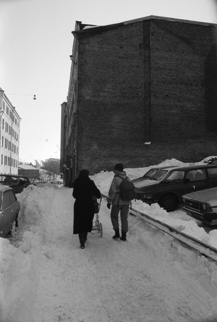 Meritullinkatu. Äiti (Etta Harvilahti) työntämässä lastenvaunuja lumisella Meritullinkadulla. Oikealla autoja Meritullinkatu 8:n rakentamattomalla tontilla. Taustalla Meritullinkatu 6:n talon tiilipääty.