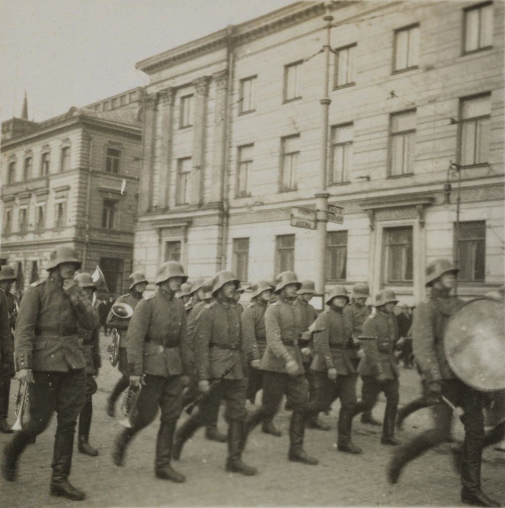 Sotilassoittokunnan paraati Helsingissä. Etualalla olevilla sotilailla on soittiminaan puhallinsoittimia, oikealla isorumpu. Taustalla Pohjoisesplanadi 15 ja Kaupungintalo.