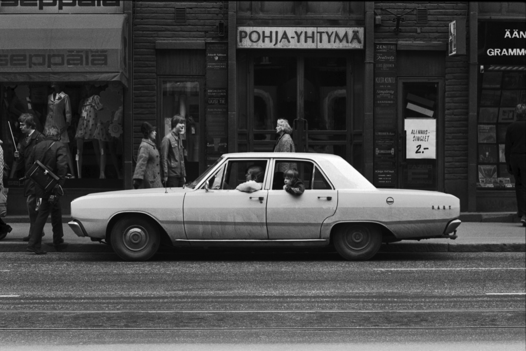 Kaisaniemenkatu 13. Nuoret miehet Kaisaniemenkatu 13:n edustalle pysäköidyssä vaaleassa autossa (Dodge Dart) seuraamassa työväen vappumarssia 1.5.1972.