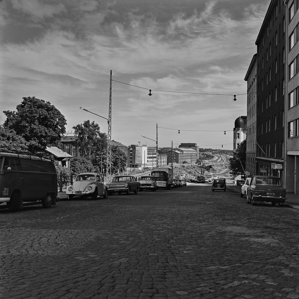 Näkymä Sörnäisistä Pääskylänkatua pitkin Kulosaaren sillan suuntaan. Oikealla Pääskylänkatu 5, 7 ja 9, vasemmalla etäämmällä Lautatarhankadun teollisuus- ja toimistokiinteistöjä. Kahden kadun välissä kulki vielä kuvanoton aikaan yksi liittymä satamarataan Hermanniin ja Toukolaan. Kesällä 1982 alkoi metroliikenne välillä Rautatieasema-Itäkeskus. Horisontissa alkaa Itäväylä. Pääskylänkatu on päällystetty katukivillä. Kuvan vasemmassa reunassa pysäköidyt ajoneuvot: Volkswagen Transporter, Volkswagen, Opel Rekord, Volga, Leyland-pakettiauto. Oikealla puolella pysäköityinä Datsun Bluebird ja Fiat 600.