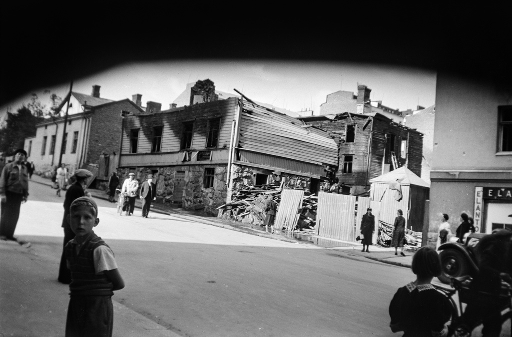 Pommitusten tuhoja Helsingissä 1941 heinäkuussa. Edessä palanut Kustaankatu 3. Etualalla vasemmalla pieni poika katsoo kameraan. Ihmiset kauhistelevat tuhoja kadulla.