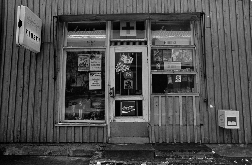 Kioski Adalmina puurakennuksessa Toukolassa. Kioskin ikkunoihin on kiinnitetty mainoksia ja lehtien lööppejä. Kioskin palveluluukun yläpuolella roikkuu myytäviä lehtiä.