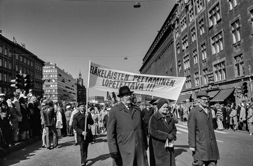 Vappumarssijoita Mannerheimintiellä Stockmannin tavaratalon kohdalla. Iäkkäät marssijat kantavat banderollia, jossa teksti: Eläkeläisten pettäminen lopetettava. Yleisö seuraa marssia kadun varrella.