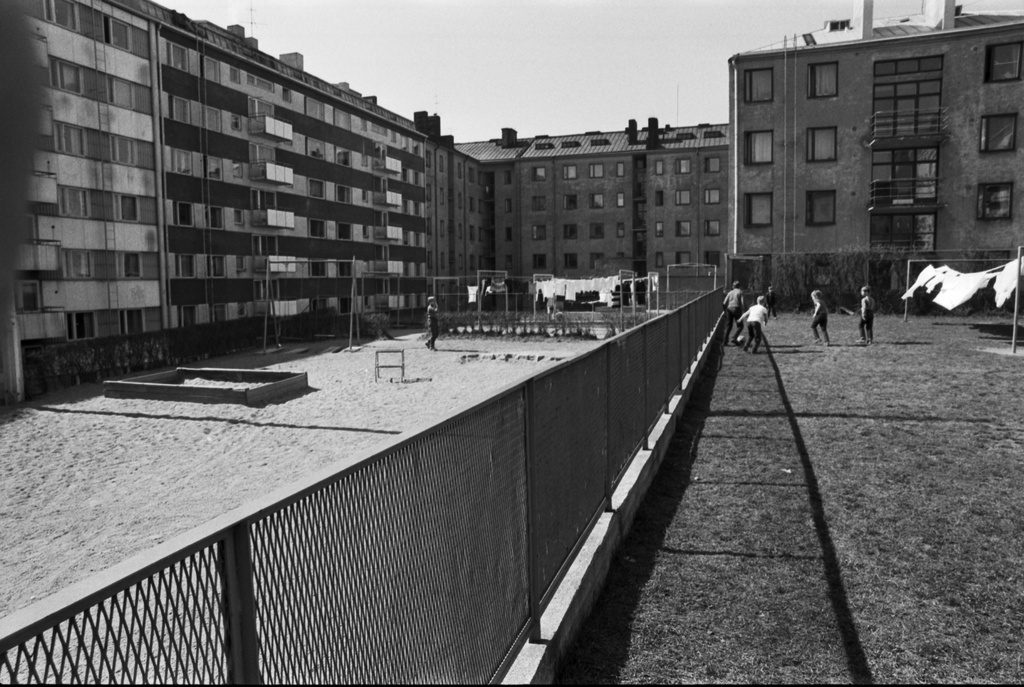 Neljäs linja 24. Näkymä Kolmas linja - Castréninkatu - Neljäs linja - Suonionkatu -katujen ympäröimän korttelin pihalta luoteeseen. Oikealla poikia pelaamassa jalkapalloa Neljäs linja 24:n nurmikkopihalla valkopyykin liehuessa narulla kuivumassa. Taaempana Neljäs linja 26 talo. Vasemmalla Kolmas linja 23 ja 25 hiekkapihaa hiekkalaatikoineen ja keinuineen. Taustalla Castréninkatu 3.
