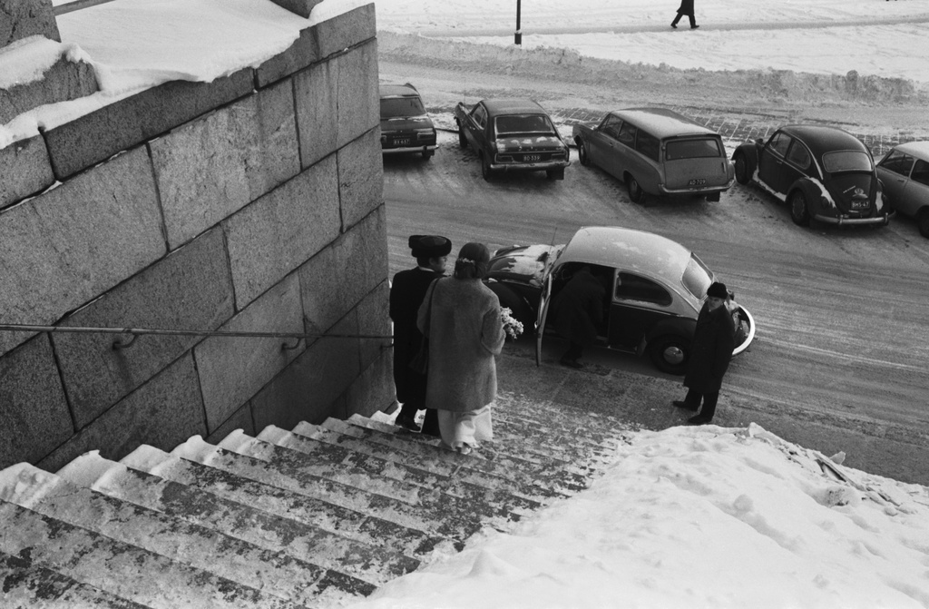 Hallituskatu 7. Hääpari kävelemässä Tuomiokirkon lumisia portaita alaspäin itäisessä kapelissa tapahtuneen vihkiäistilaisuuden jälkeen. Alhaalla odottaa auto, Volkswagen Kupla, jonka ovi avoinna. Talvi, lunta maassa.