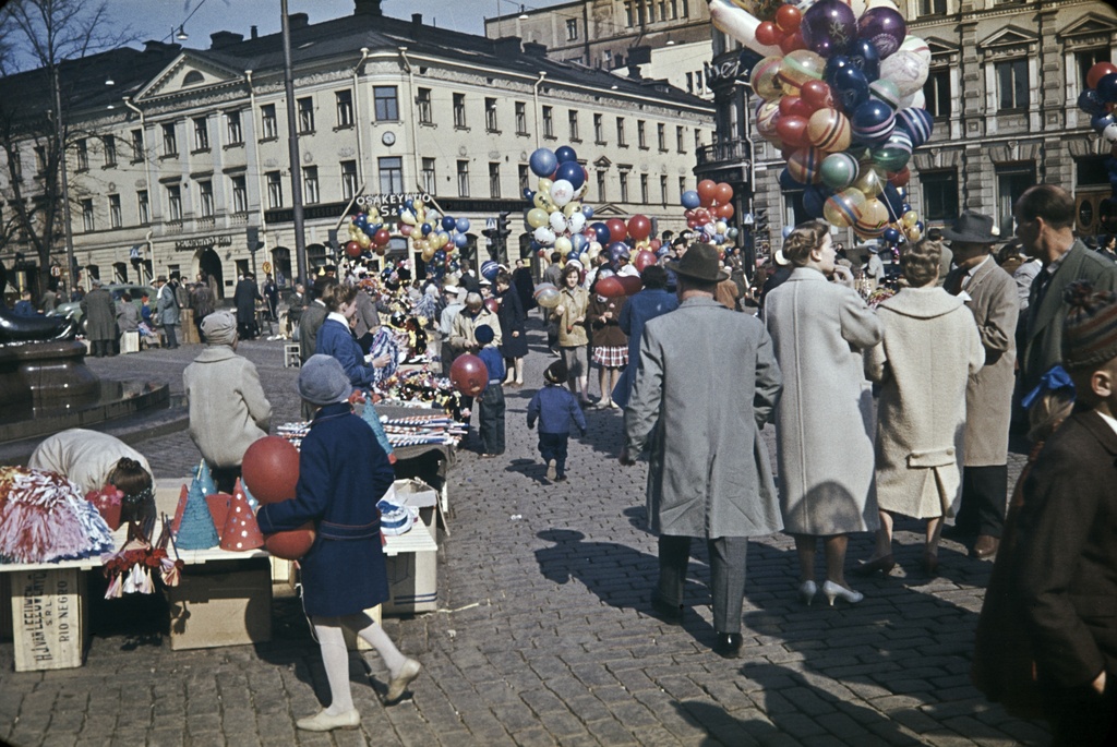 Ihmisiä Kauppatorin vapputorilla. Mm. ilmapallojen, vappuhuiskien / vappuviuhkojen ja vappuhattujen myyntiä. Taustalla Pohjoisesplanadi 17 ja 19.