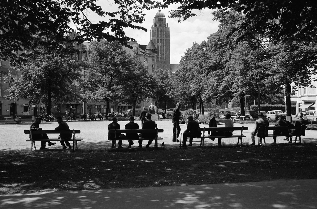 Hattupäisiä miehiä istumassa penkeillä puiden varjossa kesäisessä Karhupuistossa. Näkymä Fleminginkadun suunnalta, taustalla Kallion kirkon torni.