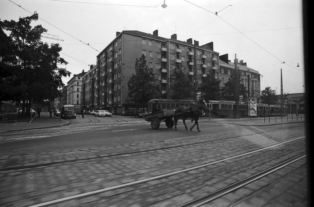 Mannerheimintie 70, 72. Mies hevosen vetämissä rattaissa ylittämässä Mannerheimintietä Sibeliuksenkadun risteyksessä. Taustalla raitiovaunuja, oikealla Töölön raitiovaunuhallit. Kuvattu ohiajavan auton ikkunasta.