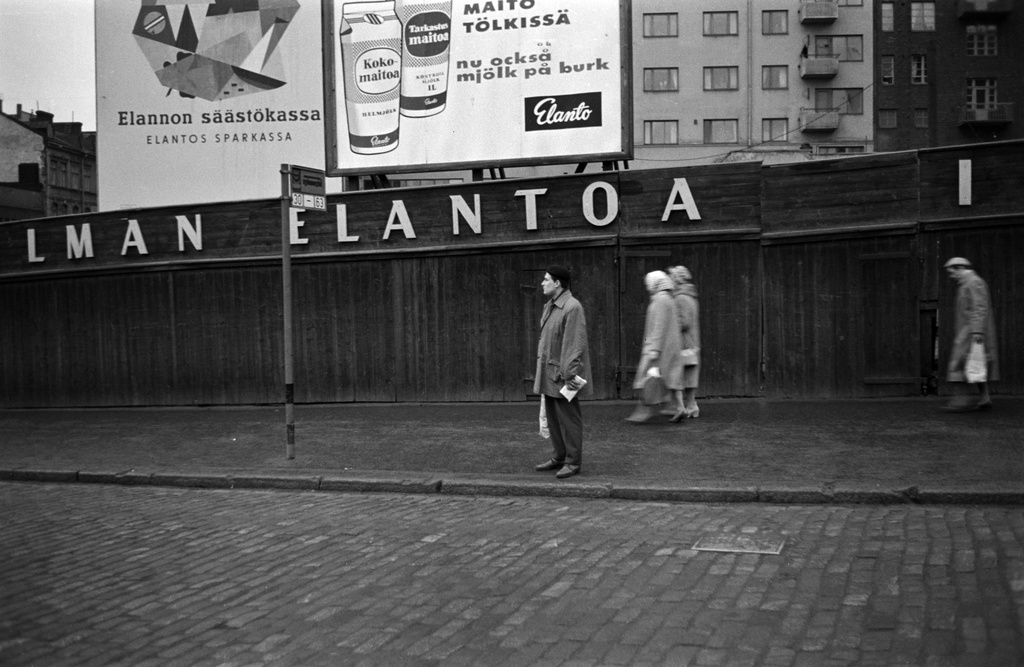 Hämeentie 16. Mies bussipysäkillä ja jalankulkijoita kadulla. Taustalla lauta-aita ja Elannon säästökassan sekä tölkissä myytävän maidon mainokset. Kuva vuodelta 1958, jolloin maitotölkit tulivat käyttöön. Hämeentie 16 tyhjä tontti. Sen takana Neljäs linja 3.