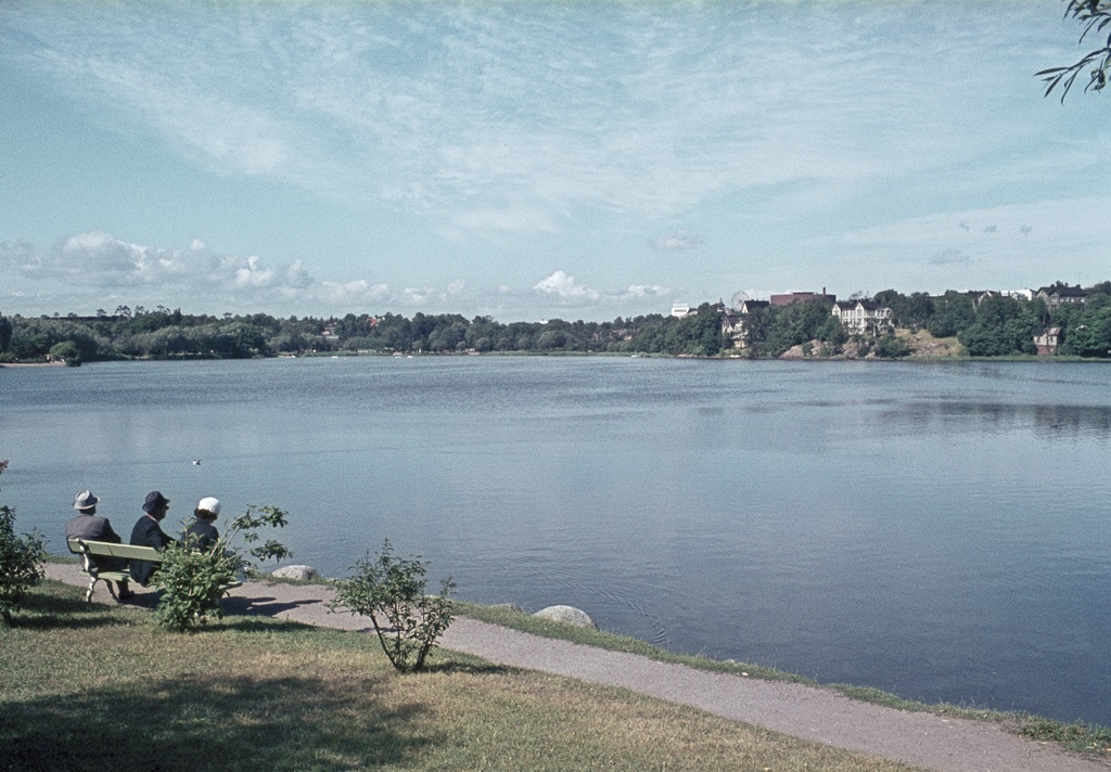 Töölönlahden rantaa, penkillä istumassa kaksi miestä ja nainen. Taustalla mm. Linnanmäki ja Linnunlaulun huviloita.