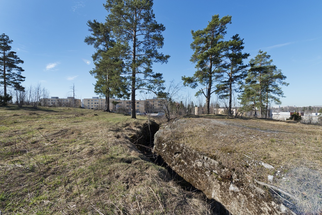 Länsimäki. Ensimmäisen maailmansodan aikainen maalinnoitus, puolustusasema II:8 Länsimäessä, Vantaalla. Kuvassa näkyy loivasti mutkittelevaa kallioon louhittua, betonilla tuettua yhdyshautaa aseman keski/itäosassa. Oikean puolen betonipuolen päällysteessä näkyy reikä. Maasto on avointa, paikoin ruohikkoista ja paikoin avokalliota. Taaempana kasvaa mäntyjä. Taustalla näkyy asuinrakennuksia sekä parkkipaikka.  Kuvaussuuntana etelä-lounas.
