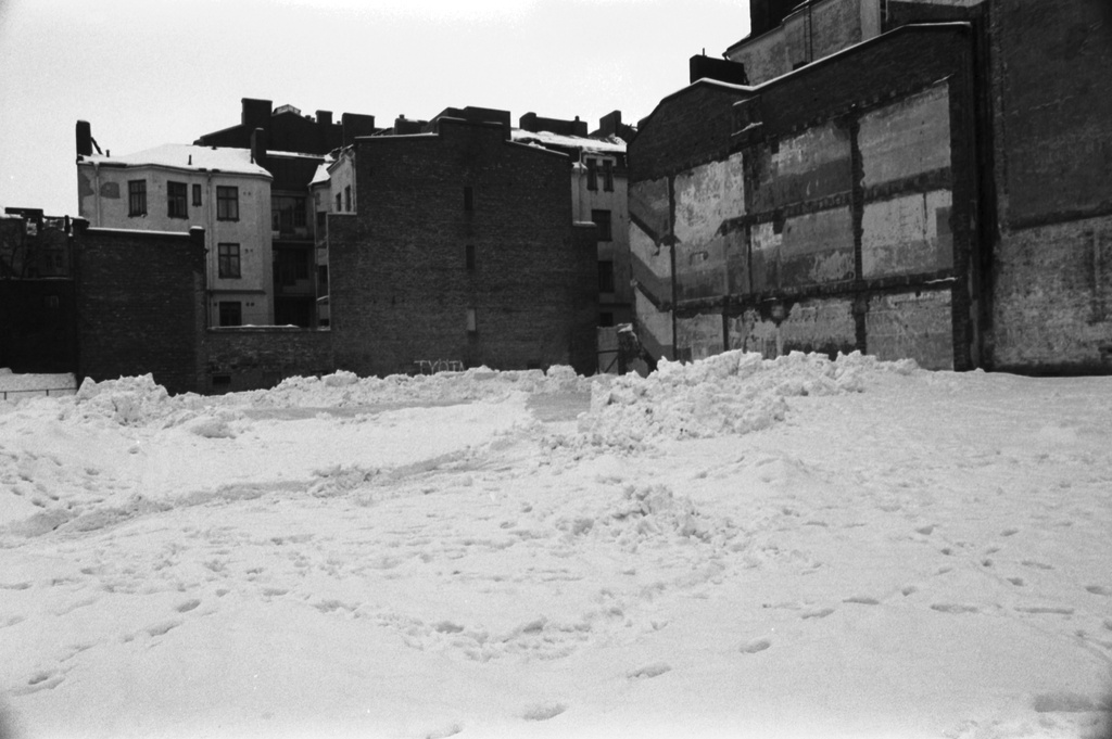 Snellmaninkatu 19. Puretun talon luminen tontti Snellmaninkatu 19:ssä. Oikealla Snellmaninkatu 17:n / Vironkatu 11:n talon seinää, jossa jälkiä tontilta puretusta talosta. Taustalla Vironkatu 9:n tontilla olevia rakennuksia.