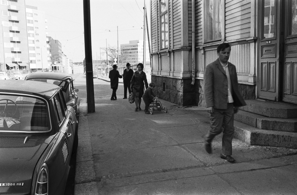 Toinen linja 8. Jalankulkijoita, kolme miestä, nainen sekä poika polkuautonsa kanssa, kävelemässä Toinen linja 6:n (nykyisin 8) puutalon edustalla. Jalkakäytävällä sementtilaatat. Näkymä Hakaniemen suuntaan.