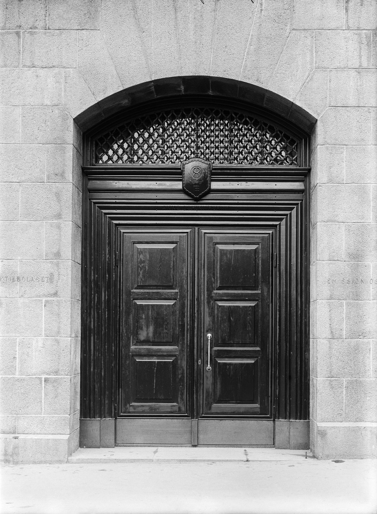 Keskuskatu 1b (Hakasalmenkatu 1). Suomen Käsityöläis-Osake-Pankin ulko-ovi. Oven yläpuolella logo, mahdollisesti Käsityöläis-Osake-Pankin liikemerkki ja teksti ANNO DOMINI MCMXXI. Vuonna 1920 valmistunut rakennus on Eliel Saarisen suunnittelema ja Käsityöläis-Osake-Pankin rakennuttama. Siinä sijaitsi pankin pääkonttori. 1910 rakennettu Kino-Palatsi sijaitsi korttelin sisällä, ja sinne pääsi Saarisen suunnitteleman talon kautta.