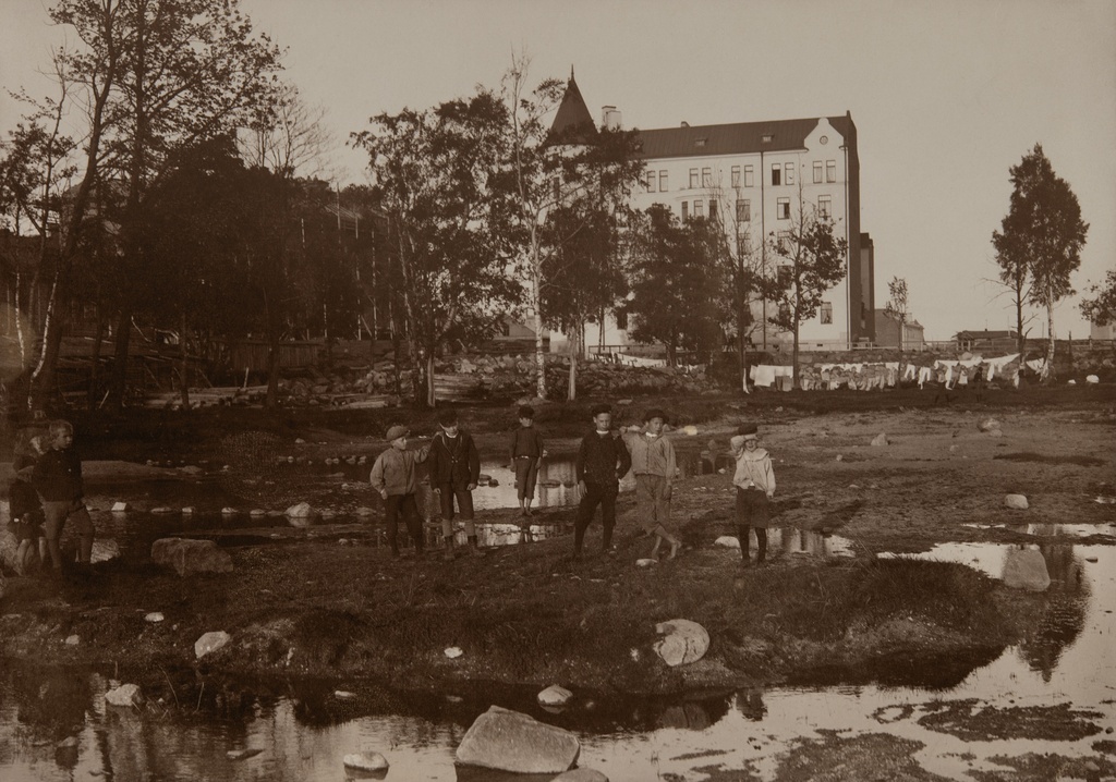 Ryhmä lapsia ja naruilla kuivamassa olevaa pyykkiä Huvilakadun ja Tehtaankadun rakentamattomalla kulmatontilla, Huvilakatu 32-30 - Tehtaankatu 30-28. Tontille rakennettiin 1905 Eiran sairaalan uudempi osa. Taustalla vuonna 1902 valmistunut asuintalo Huvilakatu 27 - Tehtaankatu 26.