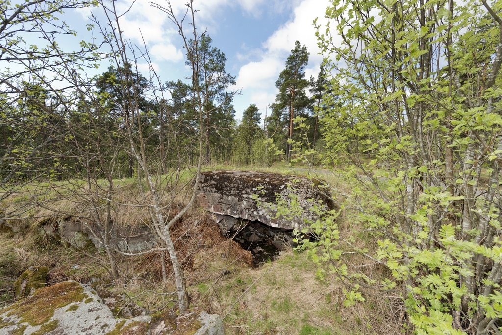 Laajalahti. Ensimmäisen maailmansodan aikainen maalinnoitus, puolustusasema XXXI:4 Laajalahdella, Espoossa. Kuvassa on muuratuista kivistä ja betonista rakennettu suojahuone. Sisäänkäynti avautuu kuvassa vasemmalle jatkuvaan, kallioon louhittuun yhdyshautaan, jota on tuettu betonilla sekä harkkokivimuurauksella. Huoneen lattialla on muurikiviä ja roskia. Ympäristössä kasvaa nuorta lehtipuustoa sekä mäntyvaltaista metsää.
