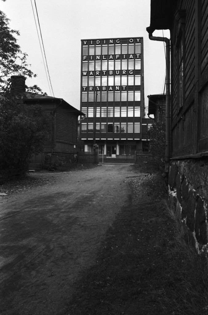 Näkymä Ruoholahden villojen pihapiiristä pohjoiseen. Taustalla Lastenkodinkatu 5 toimistotalon pääty, jossa tekstit Viding Oy, Finlafiat, Wartburg sekä Trabant.